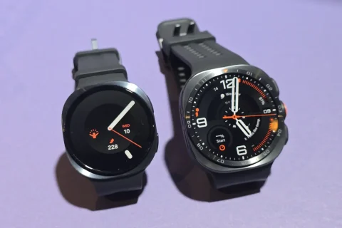 Two smartwatches on a purple surface: a round-faced watch on the left with white/orange hands, and a larger multi-dial sports watch on the right with orange accents and black strap.