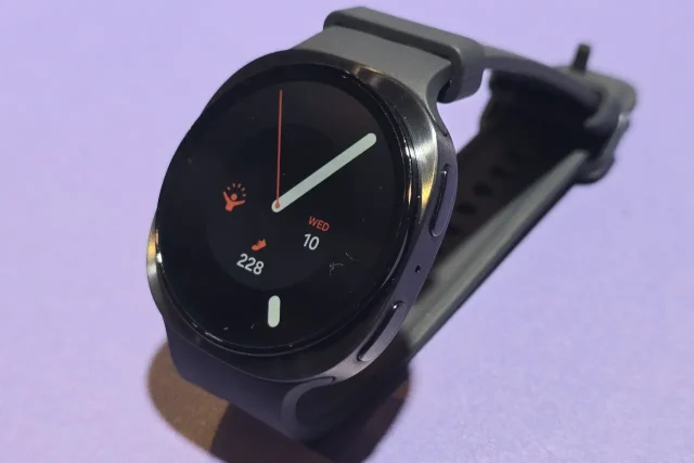 Round smartwatch on a gray band resting on a purple surface, displaying time and fitness stats (steps, date)./ Round smartwatch with circular display showing time and activity metrics on a black background.