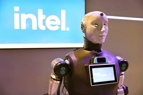 Humanoid robot with a touchscreen panel on its chest standing in front of an Intel sign at a tech event, purple lighting.